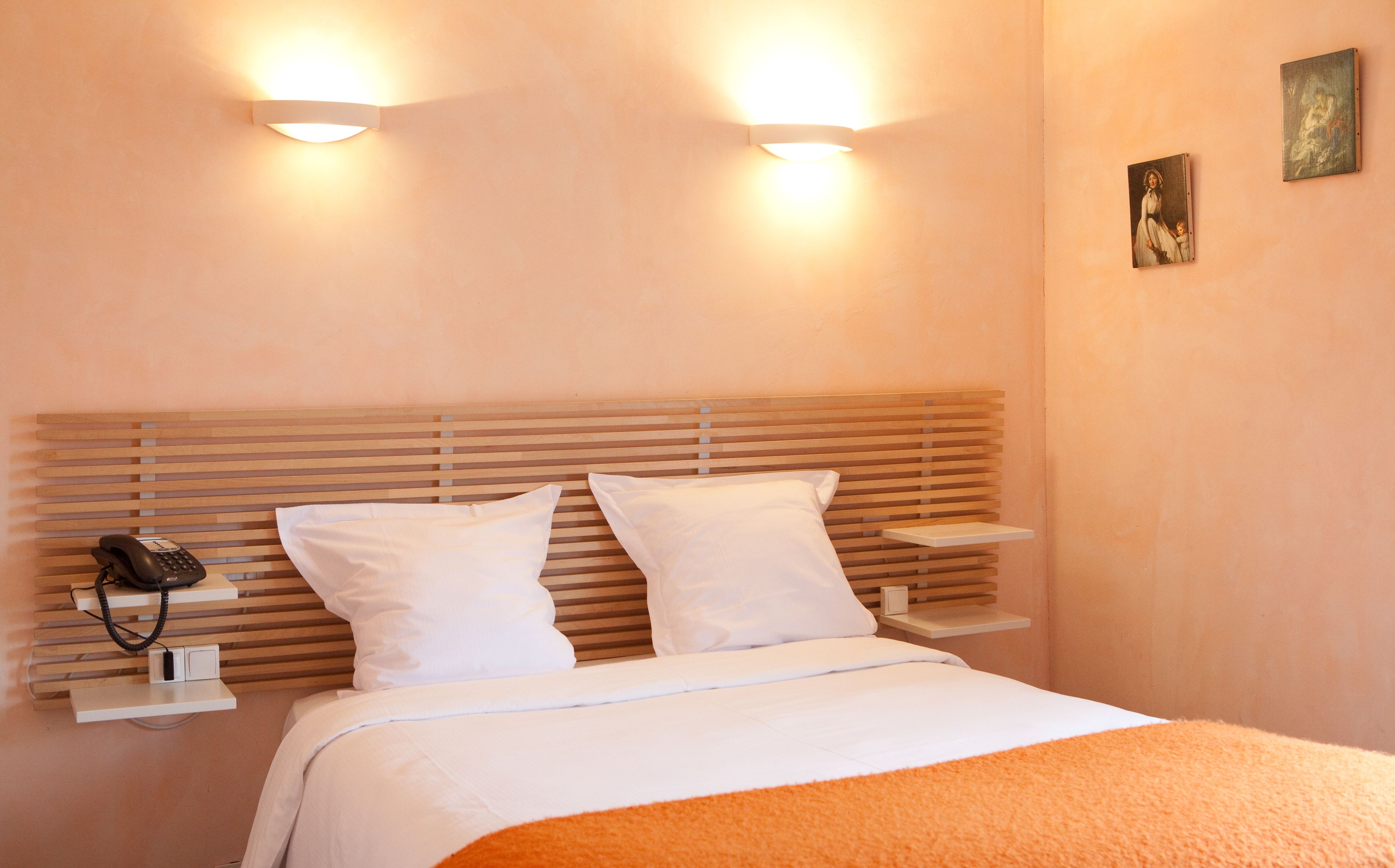 A neatly made bed with white bedding and an orange throw blanket, two white pillows, and a wooden headboard. There are two bedside tables with a phone and a small lamp on each.