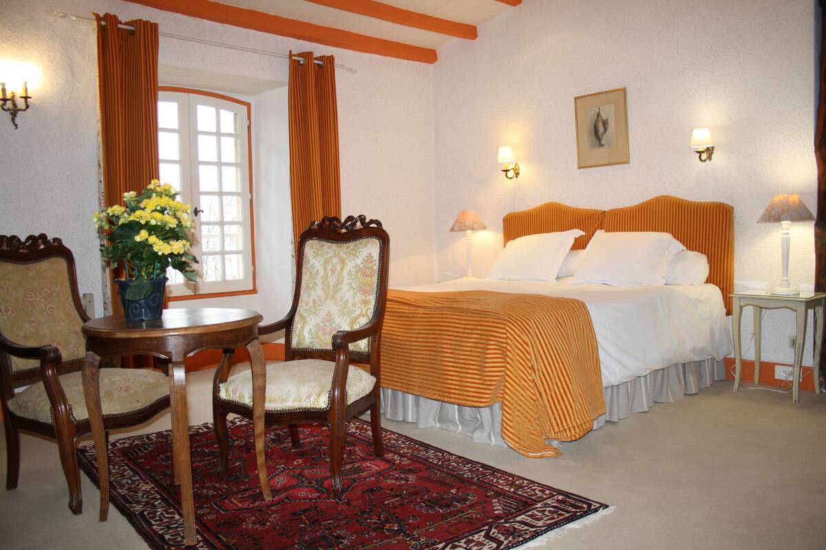 A hotel room with a bed, two chairs, a table with a vase of flowers, a red patterned rug, and two wall-mounted lamps.