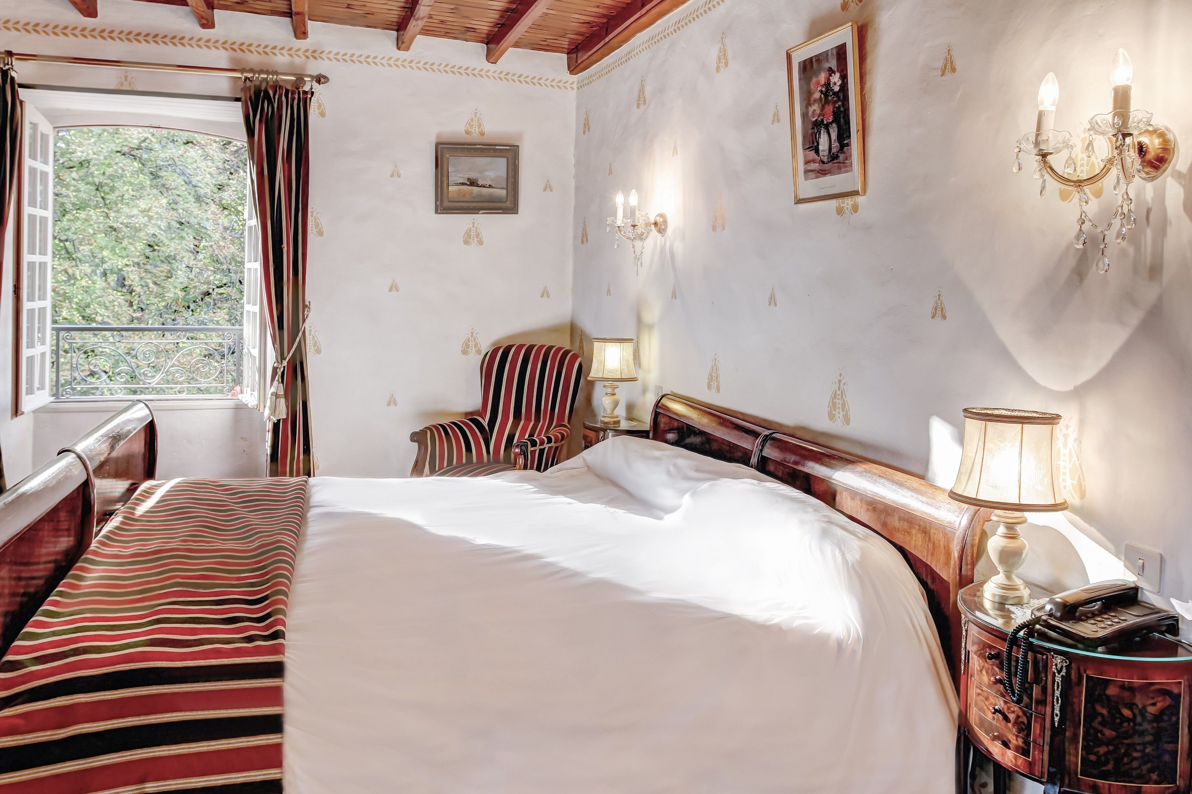 Habitación de hotel con una cama grande, una silla a rayas, una mesita de noche con lámpara y vistas a zonas verdes.