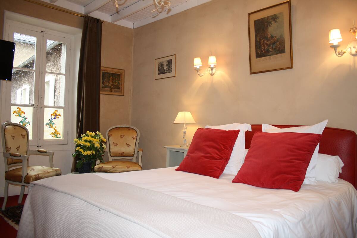 A hotel room with a bed, two chairs, a table, a lamp, and wall-mounted lights.