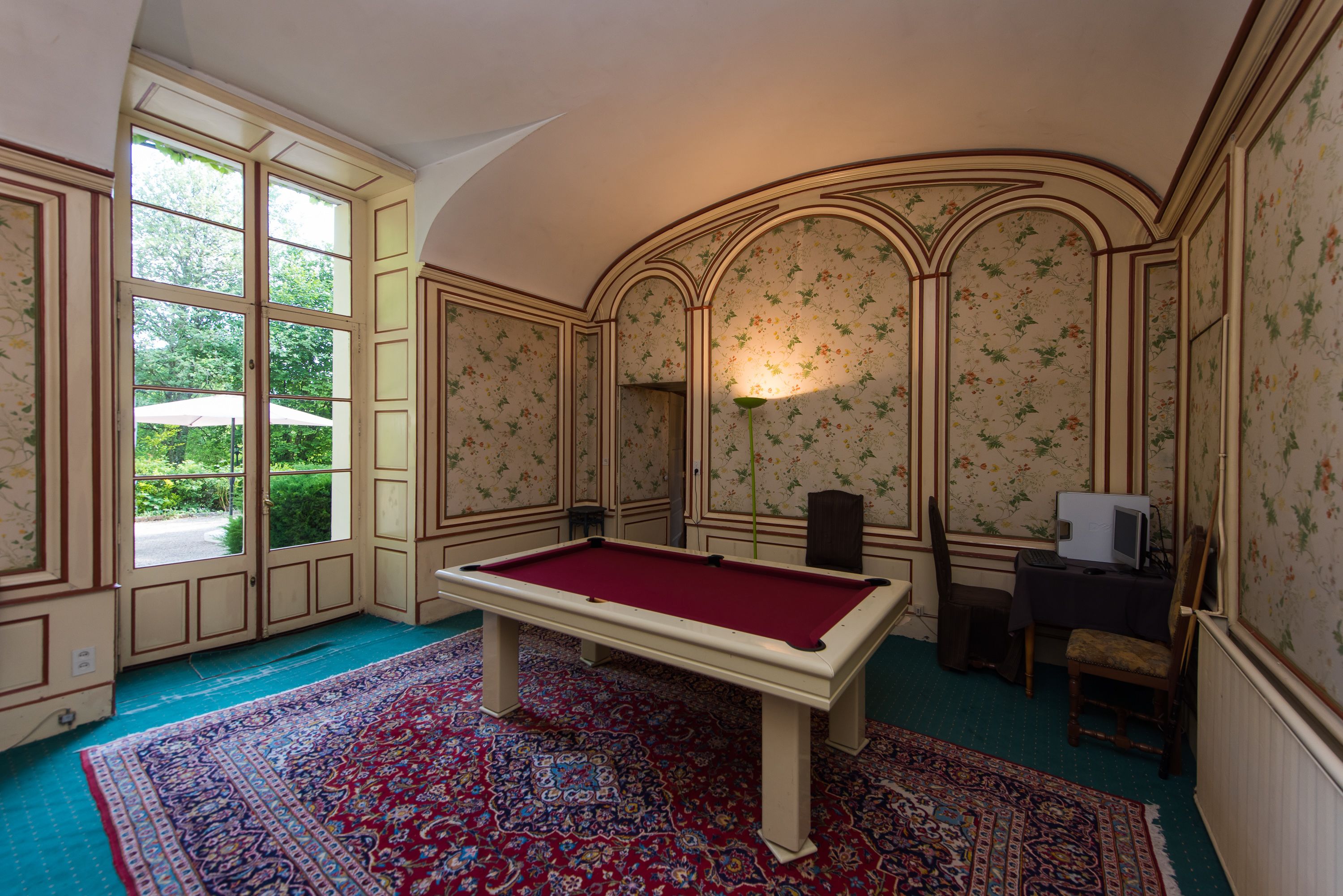 Ein Billardtisch in einem Raum mit Blumenmuster-Tapete, einem großen Fenster und einem gemusterten Teppich.
