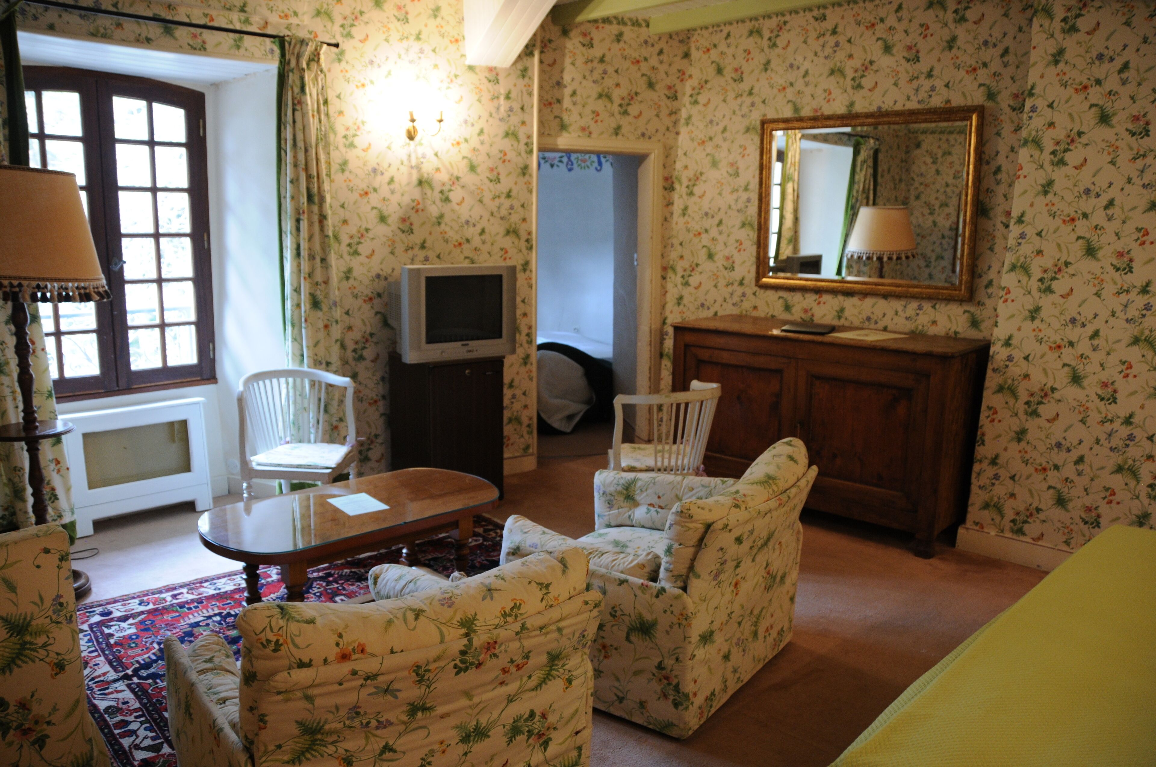 A room with floral wallpaper, a wooden cabinet, a television, a mirror, and a window with curtains.