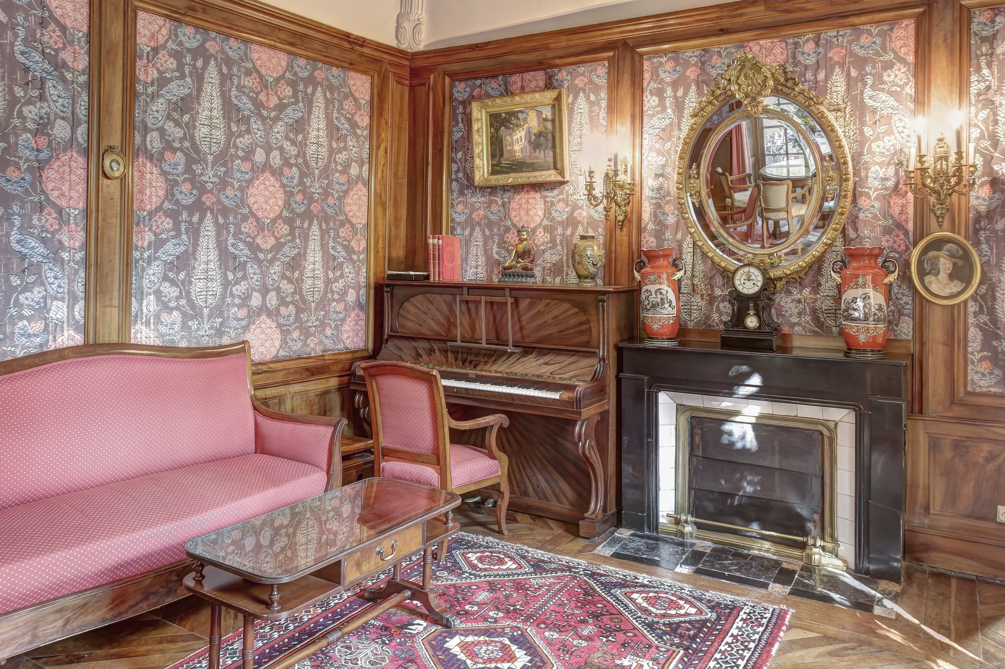 A room with a pink sofa, a wooden piano, a fireplace, and ornate wallpaper.