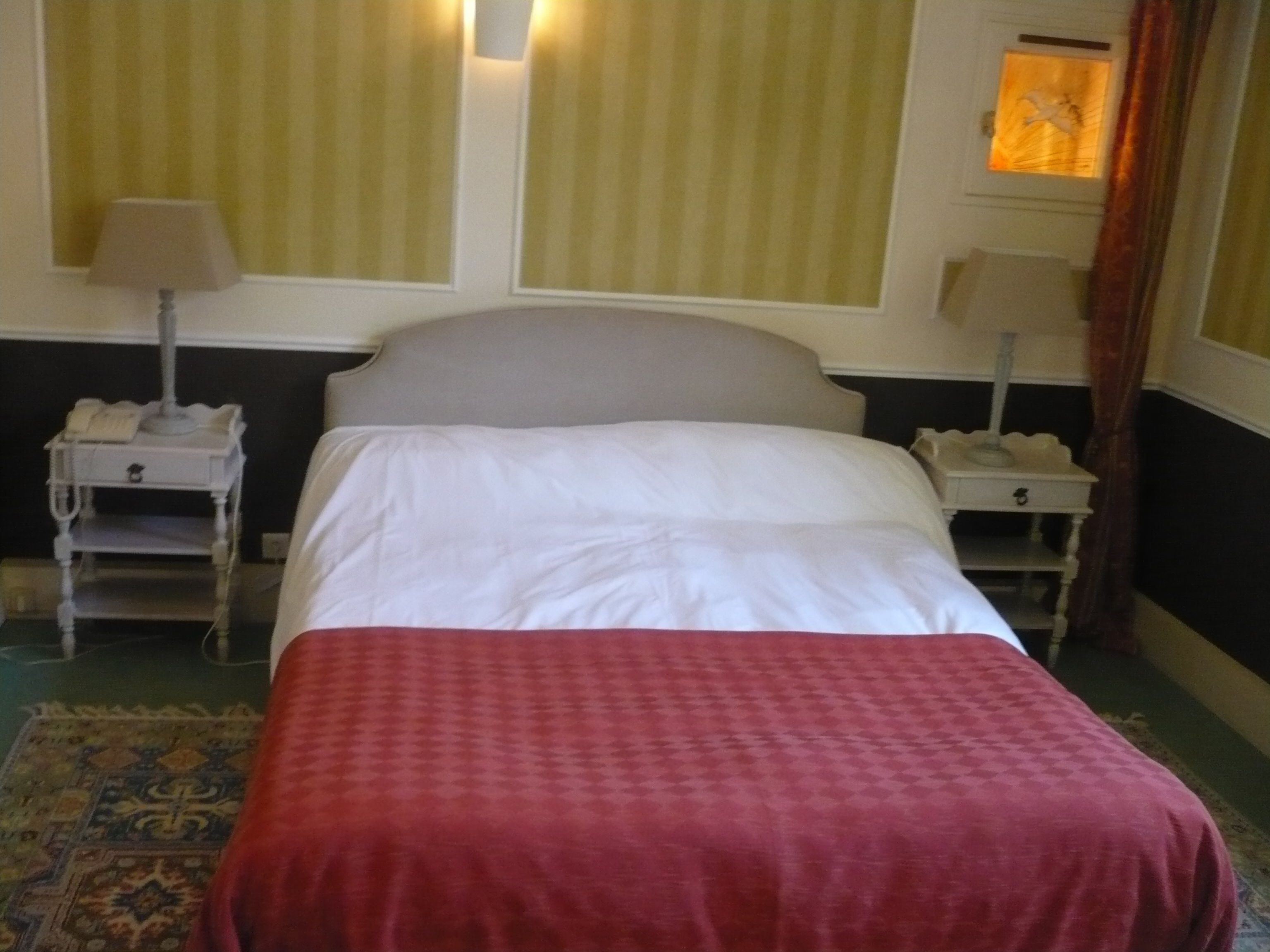 Una habitación de hotel con una cama, dos mesitas de noche, una lámpara, un teléfono y una ventana con cortinas.