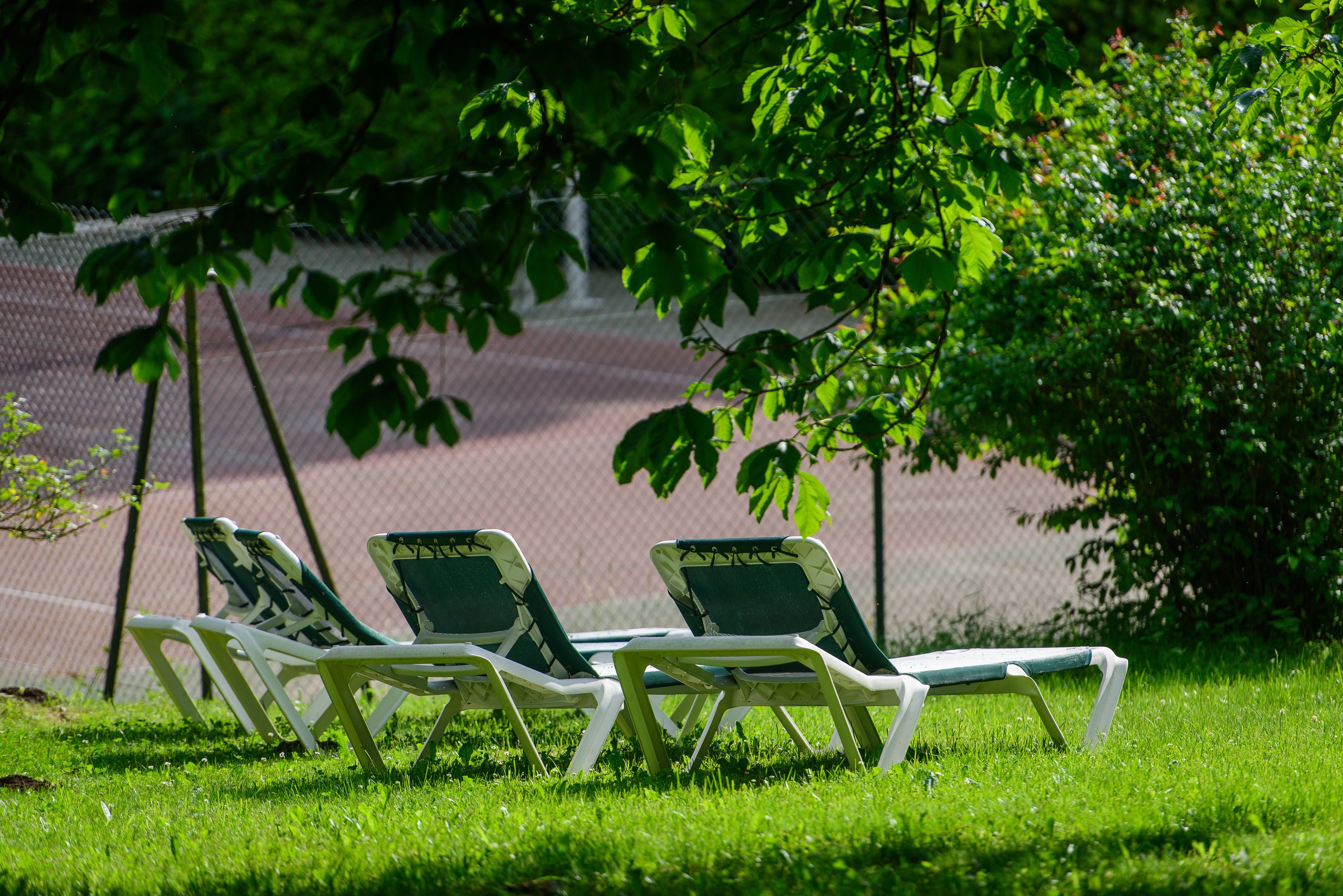Three white lounge chairs with green cushions on a grassy area near a tennis court.
