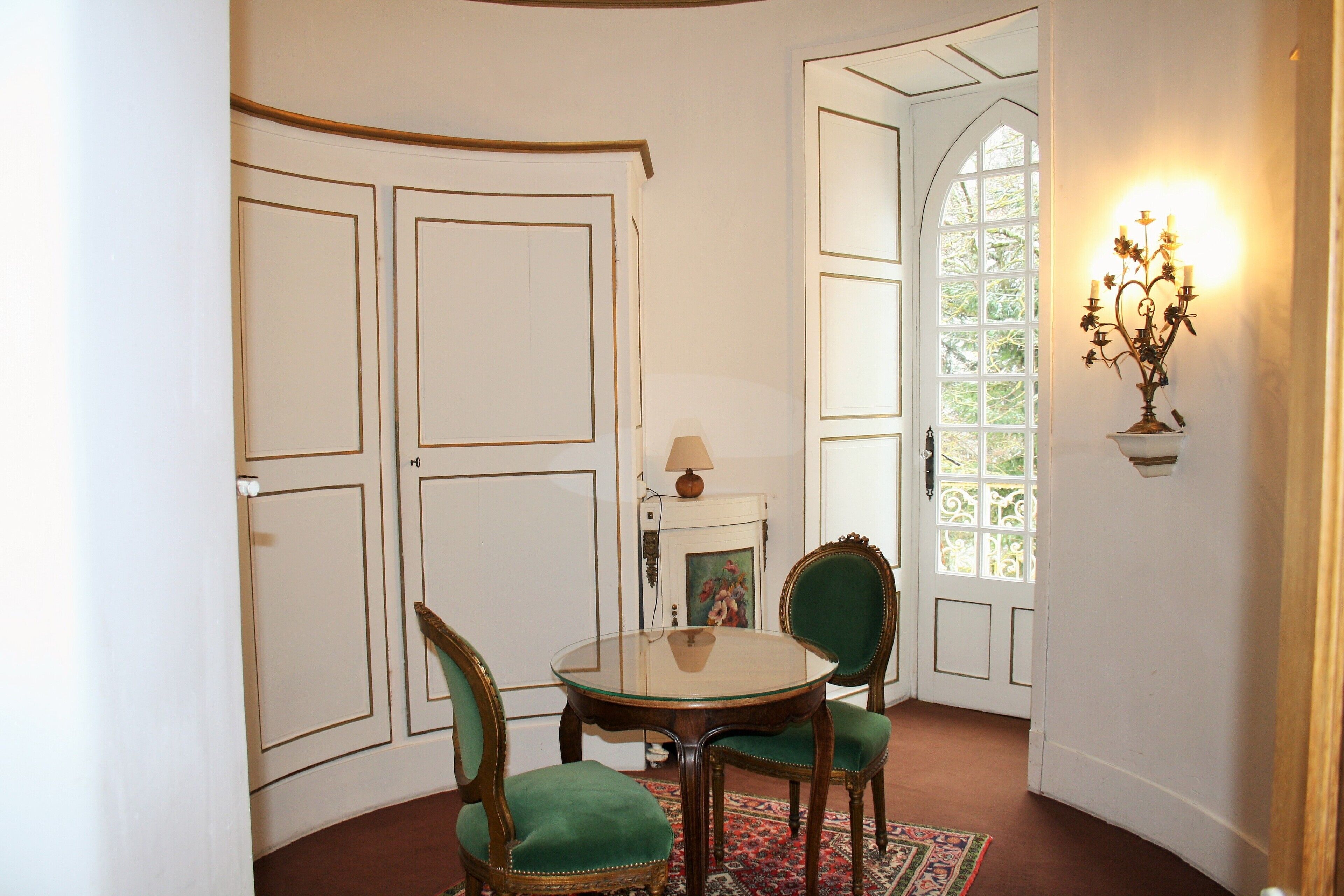 A room with a round table, two green upholstered chairs, a patterned rug, and a wall-mounted candelabra.