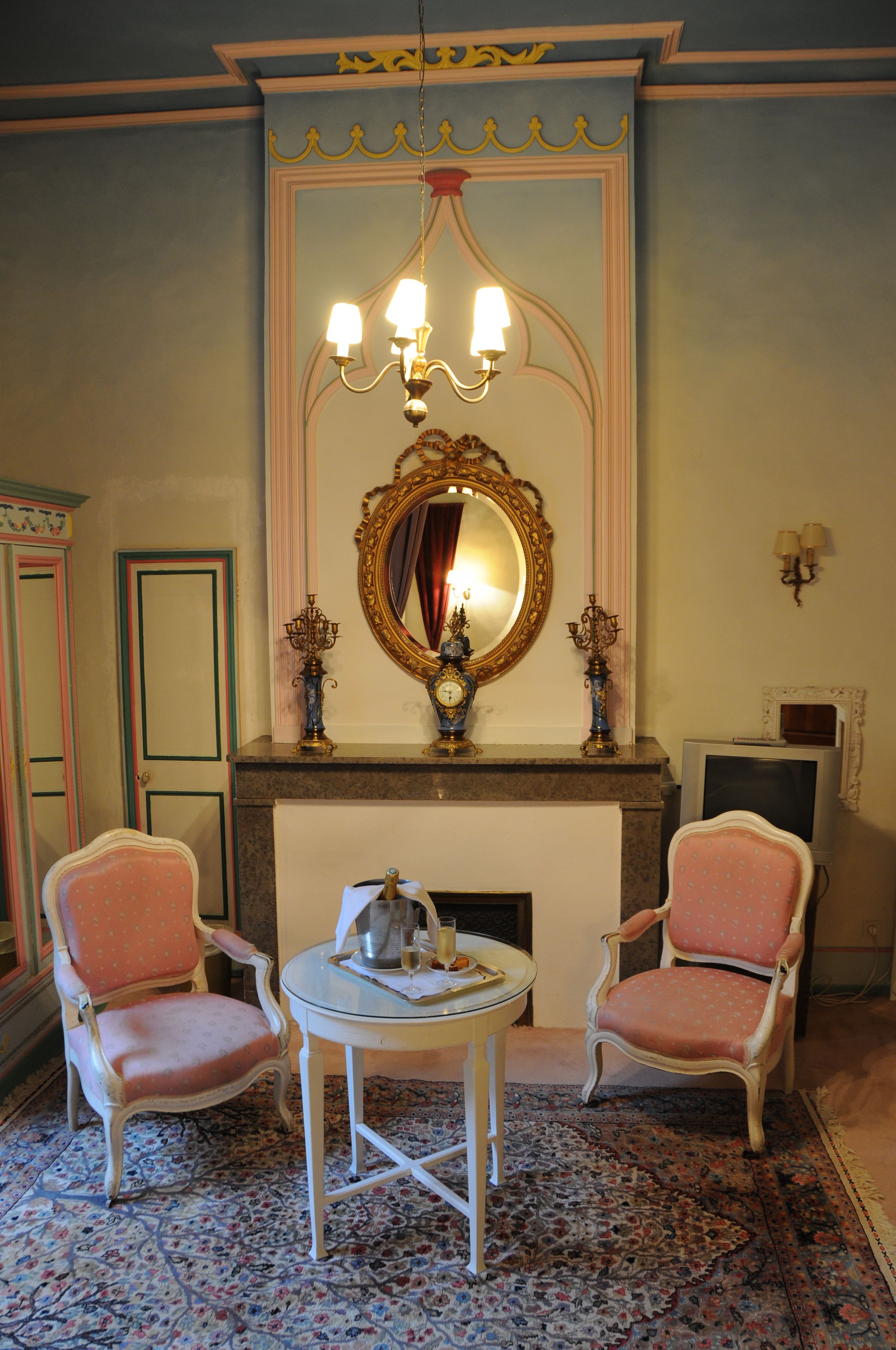 A room with a fireplace, two chairs, a small table, a mirror, and a television.