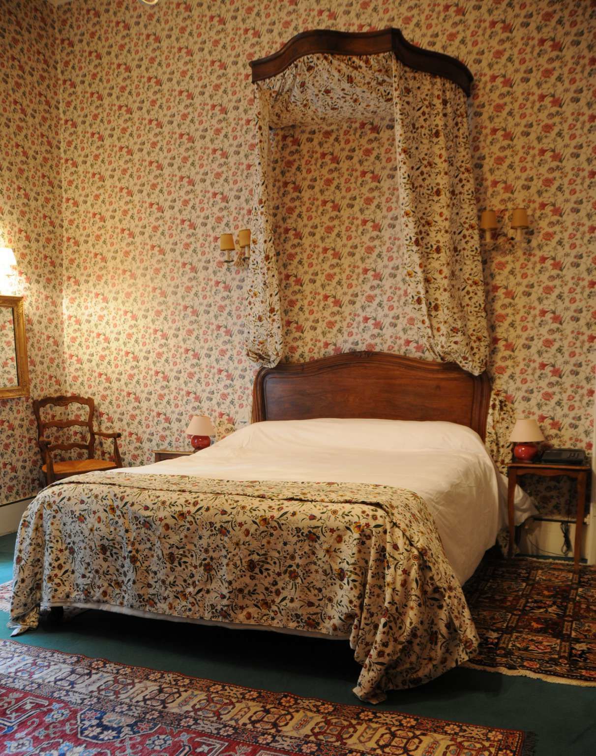 A bedroom with a four-poster bed, floral wallpaper, a patterned rug, and a nightstand with a lamp.