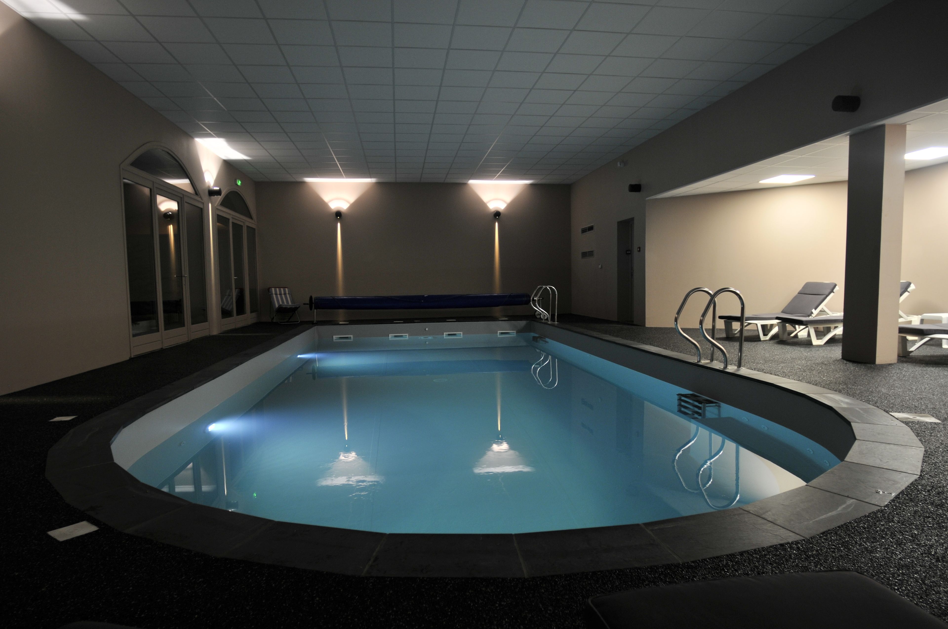 Une piscine intérieure à l’eau claire, entourée d’un sol et de murs carrelés, avec un banc et des chaises longues le long du bord.