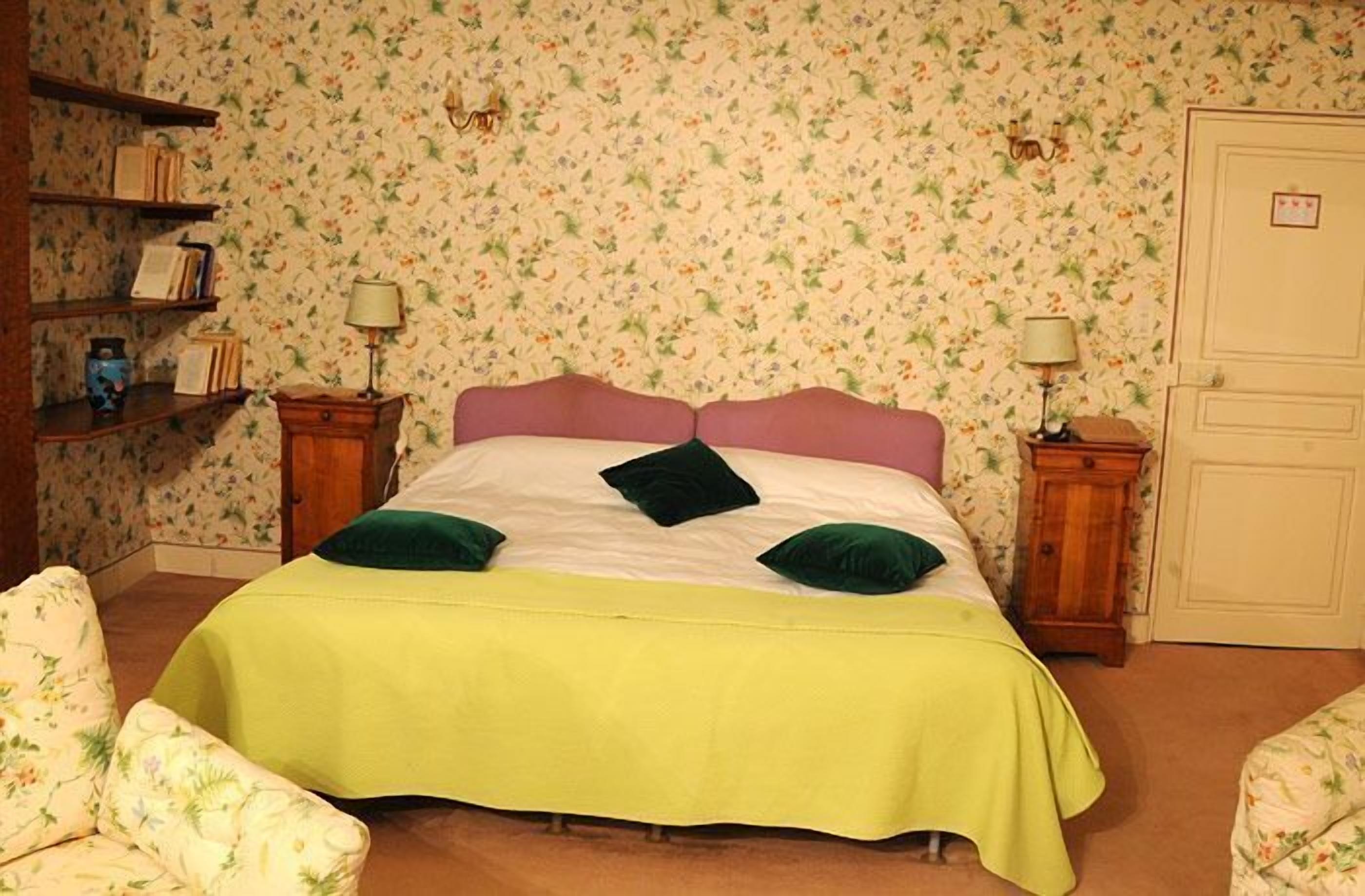 Une chambre avec un papier peint à motifs floraux, un lit recouvert d’une couverture verte, deux oreillers verts, une table de chevet en bois avec une lampe, et une chaise avec un revêtement floral.