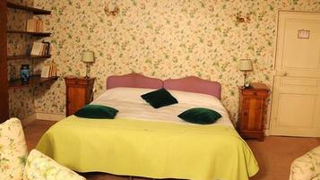 Une chambre avec un papier peint à motifs floraux, un lit recouvert d’une couverture verte, deux oreillers verts, une table de chevet en bois avec une lampe, et une chaise avec un revêtement floral.