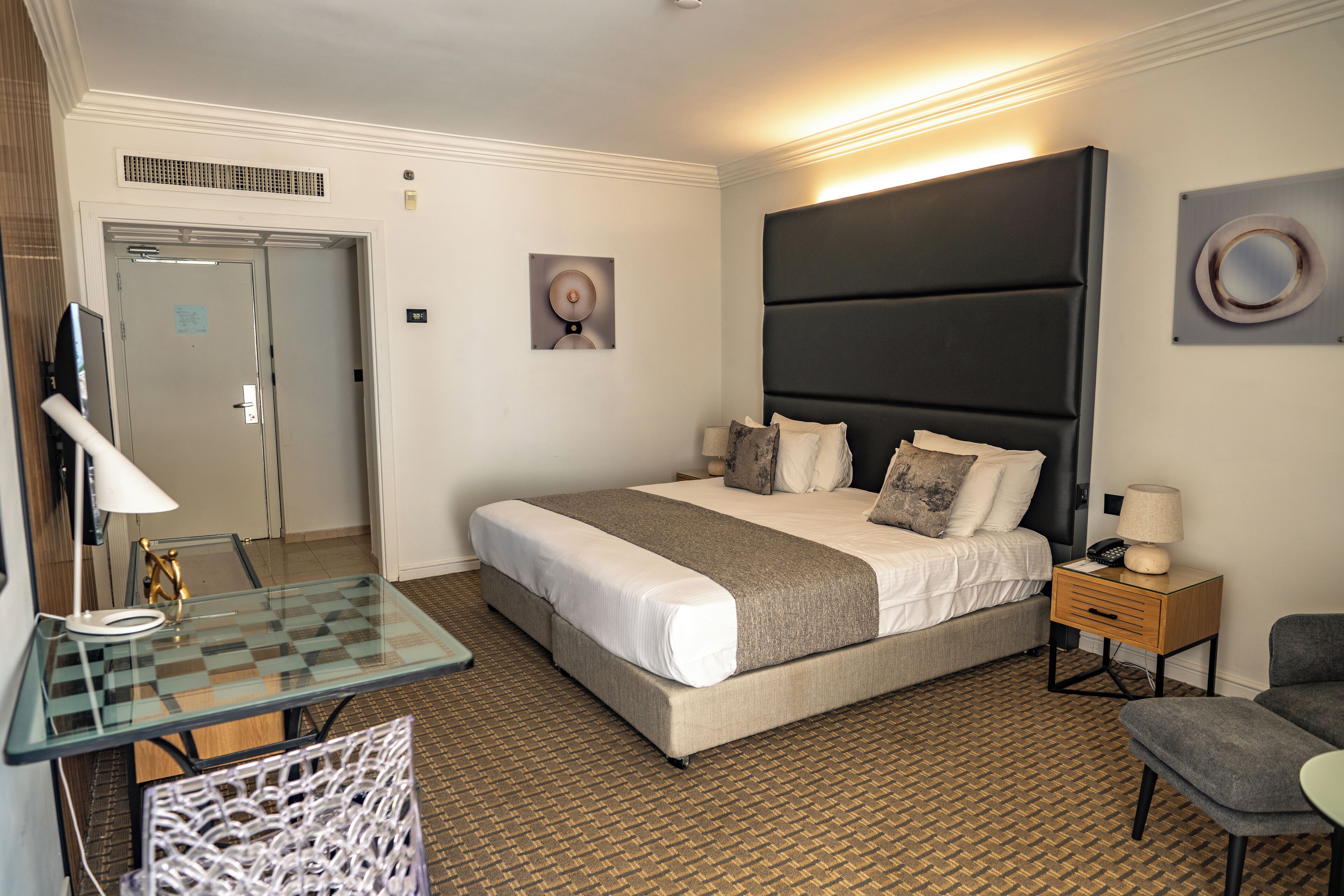 A hotel room with a bed, a glass table, a chair, a bedside table with a lamp, and a wall-mounted artwork.