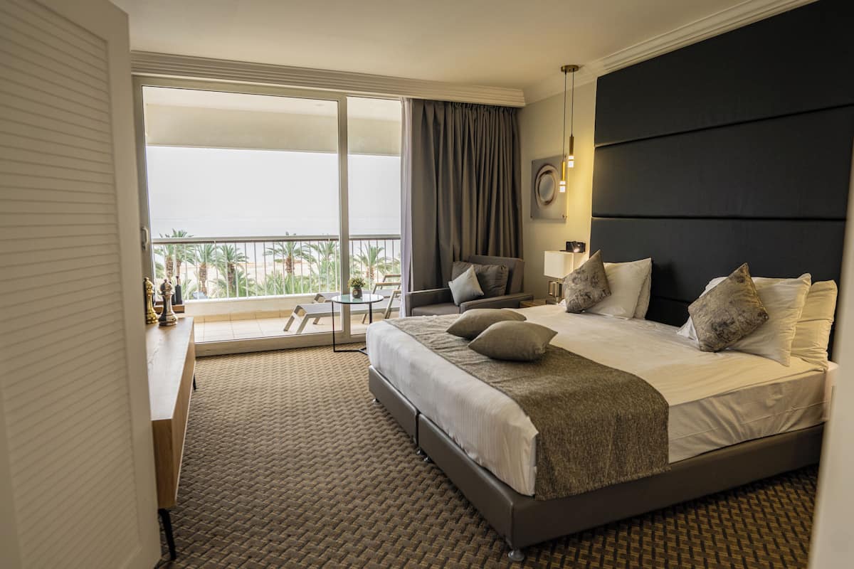 A hotel room with a large bed, a desk, a chair, a balcony with a view, and a dark headboard.