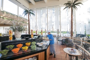 A buffet area with various food options, a waiter, and a tropical decor with palm trees and large windows. - David Dead Sea Resort & Spa (Tamar)