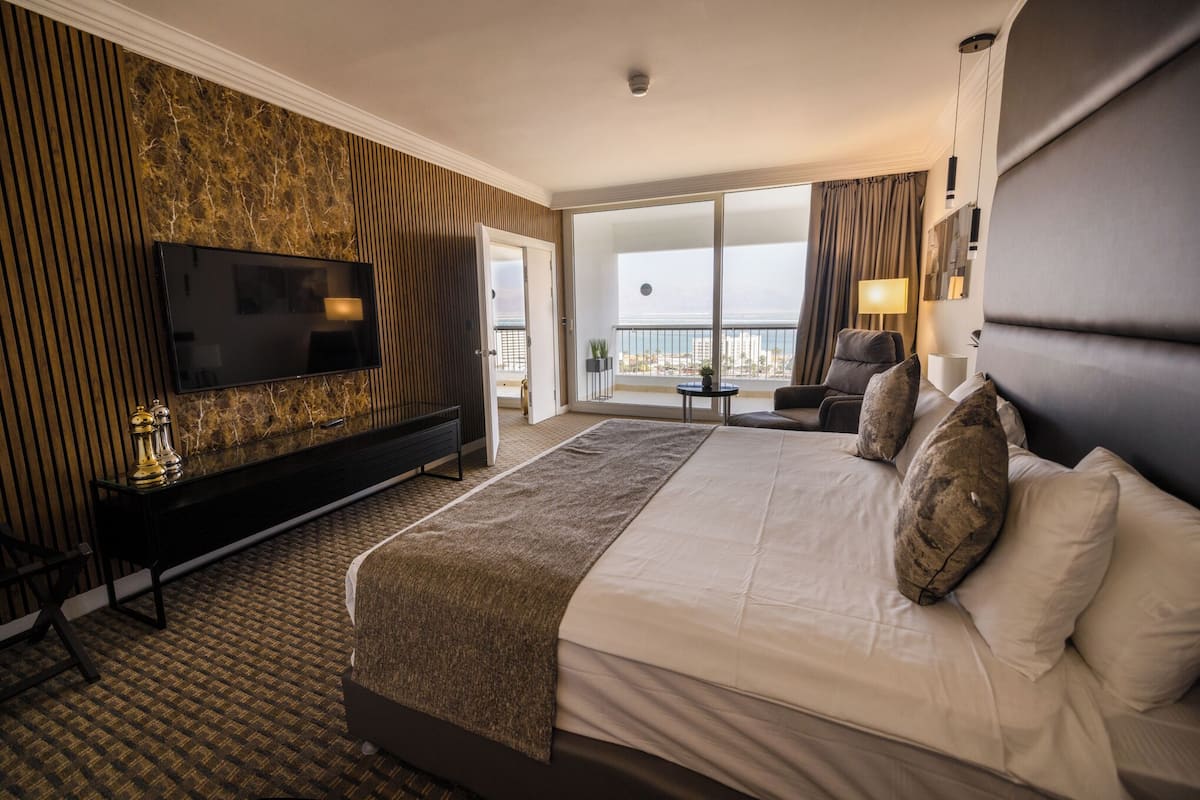 A hotel room with a large bed, a TV mounted on a marble wall, a balcony with a view, and a small table with a lamp.