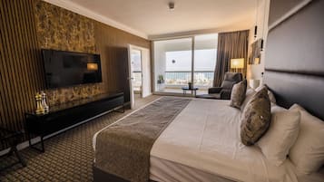 A hotel room with a large bed, a TV mounted on a marble wall, a balcony with a view, and a small table with a lamp.