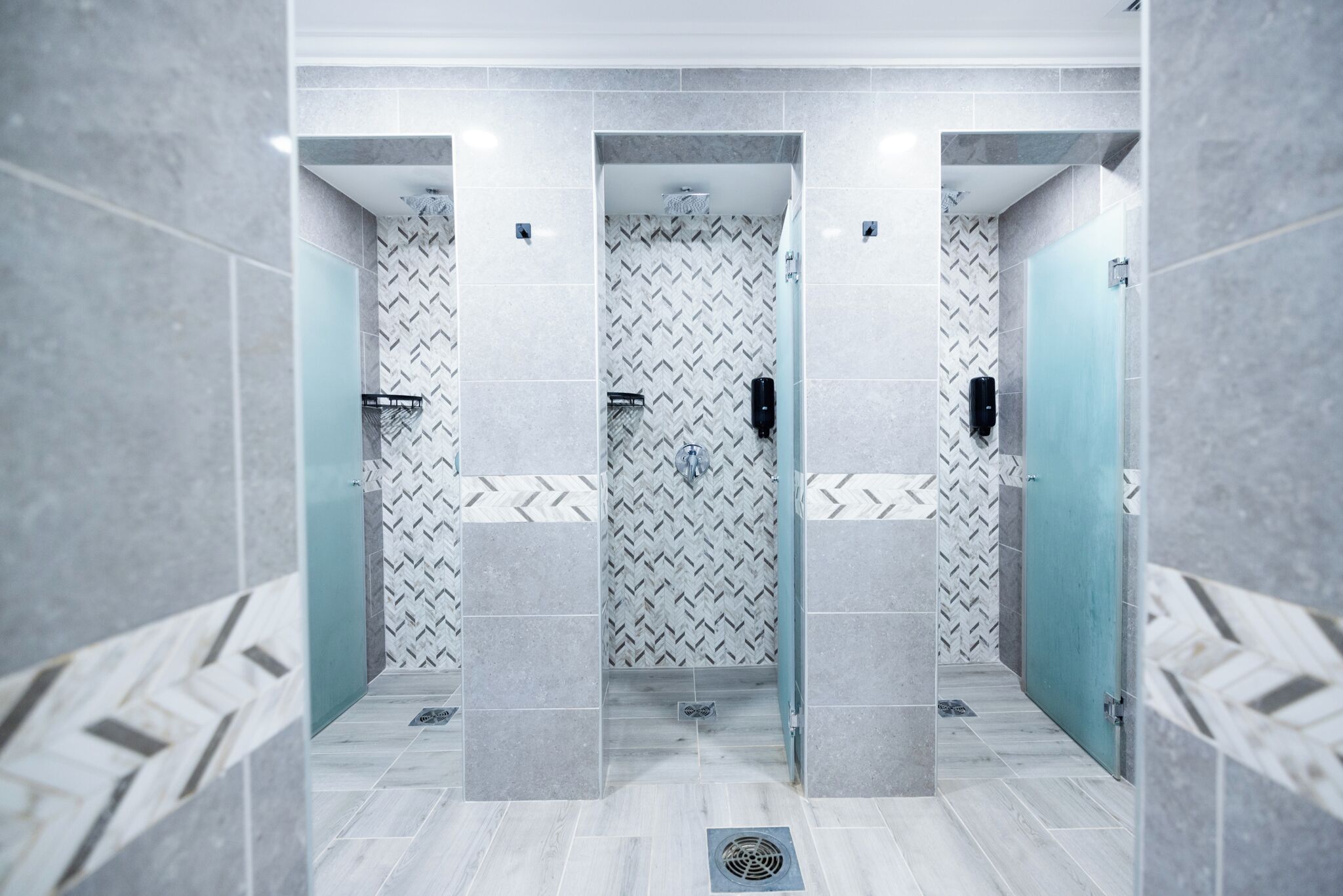 Un baño moderno con un patrón de baldosas en espiga, puertas de ducha de vidrio esmerilado y una rejilla de ventilación metálica en el suelo.