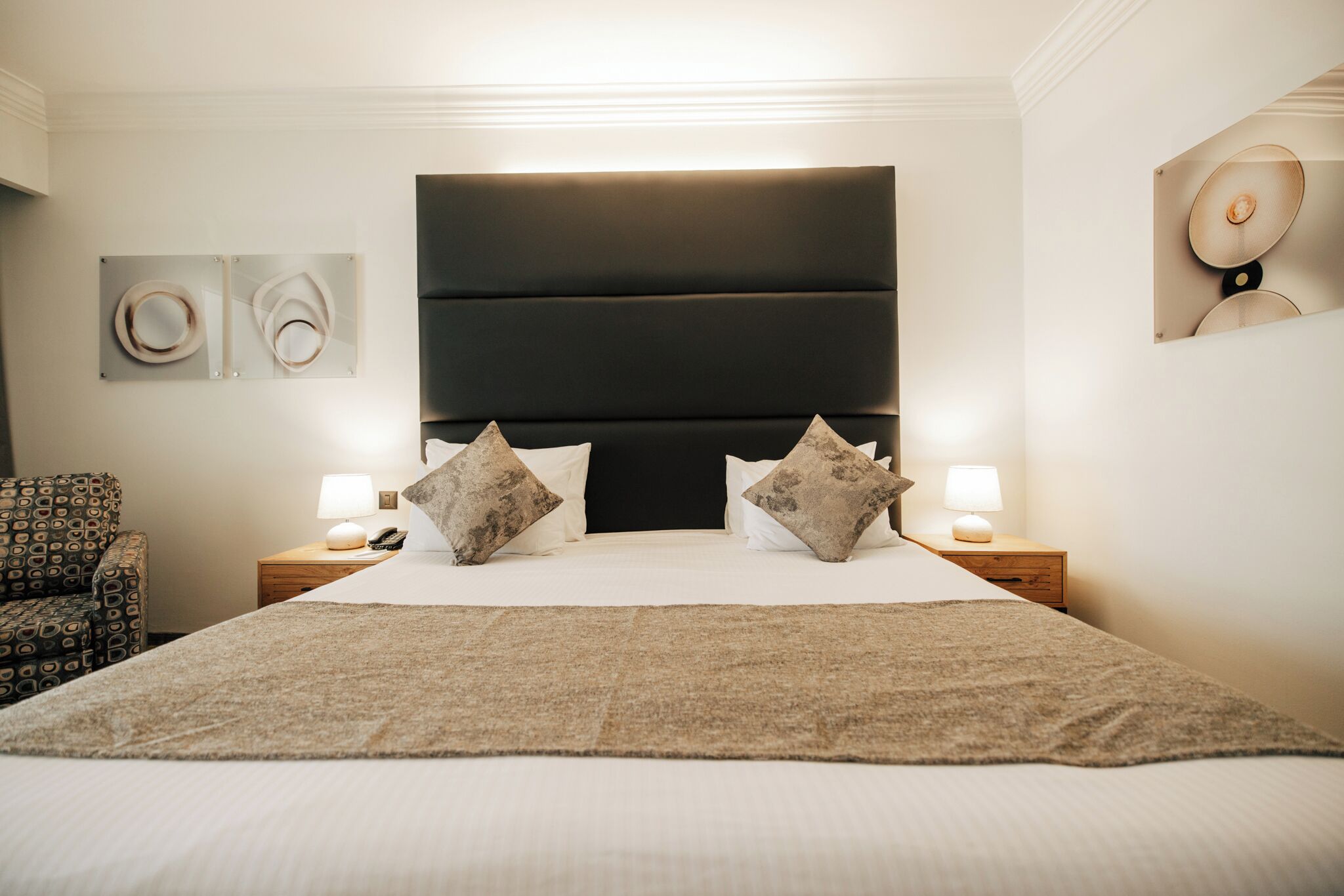 A hotel room with a large bed, two bedside tables with lamps, a patterned armchair, and artwork on the walls.