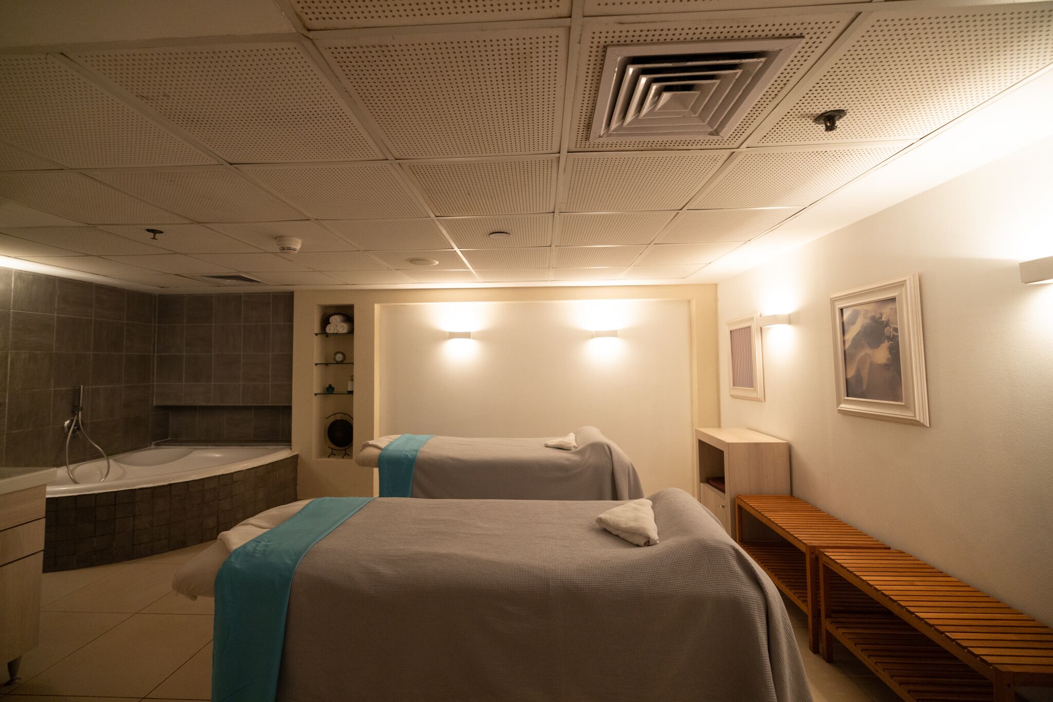 A hotel room with two beds, a bathtub, a wooden bench, and a wall-mounted shelf.