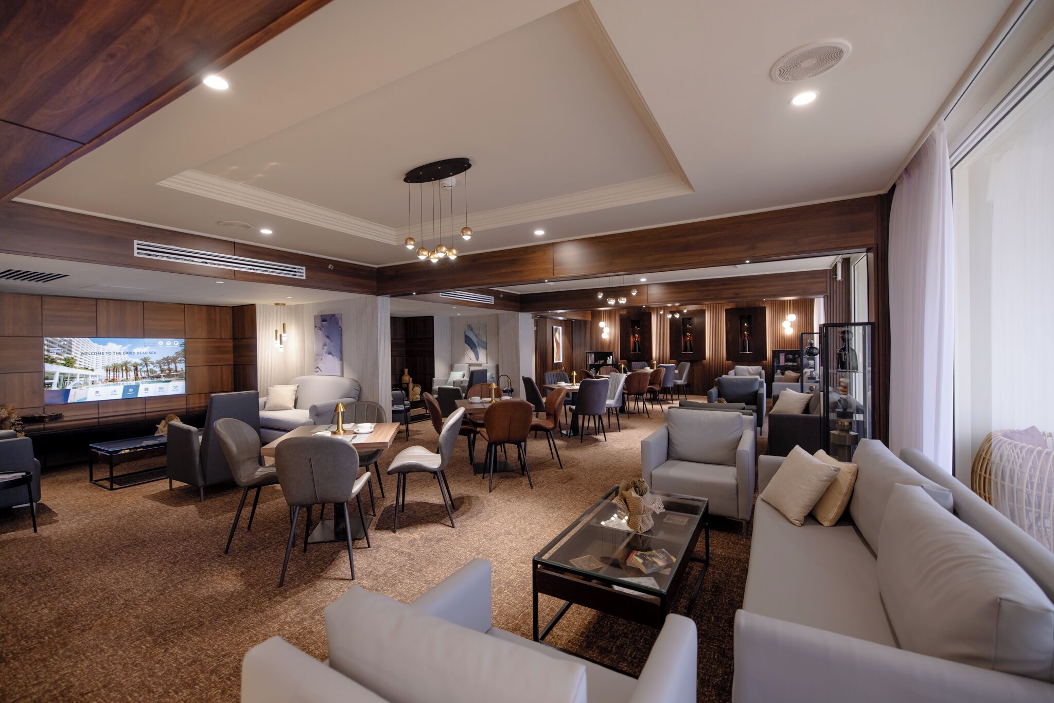 Um hall de hotel espaçoso com design moderno, apresentando uma ampla área de estar com sofás e poltronas, uma tela de televisão e um teto bem iluminado com iluminação embutida.