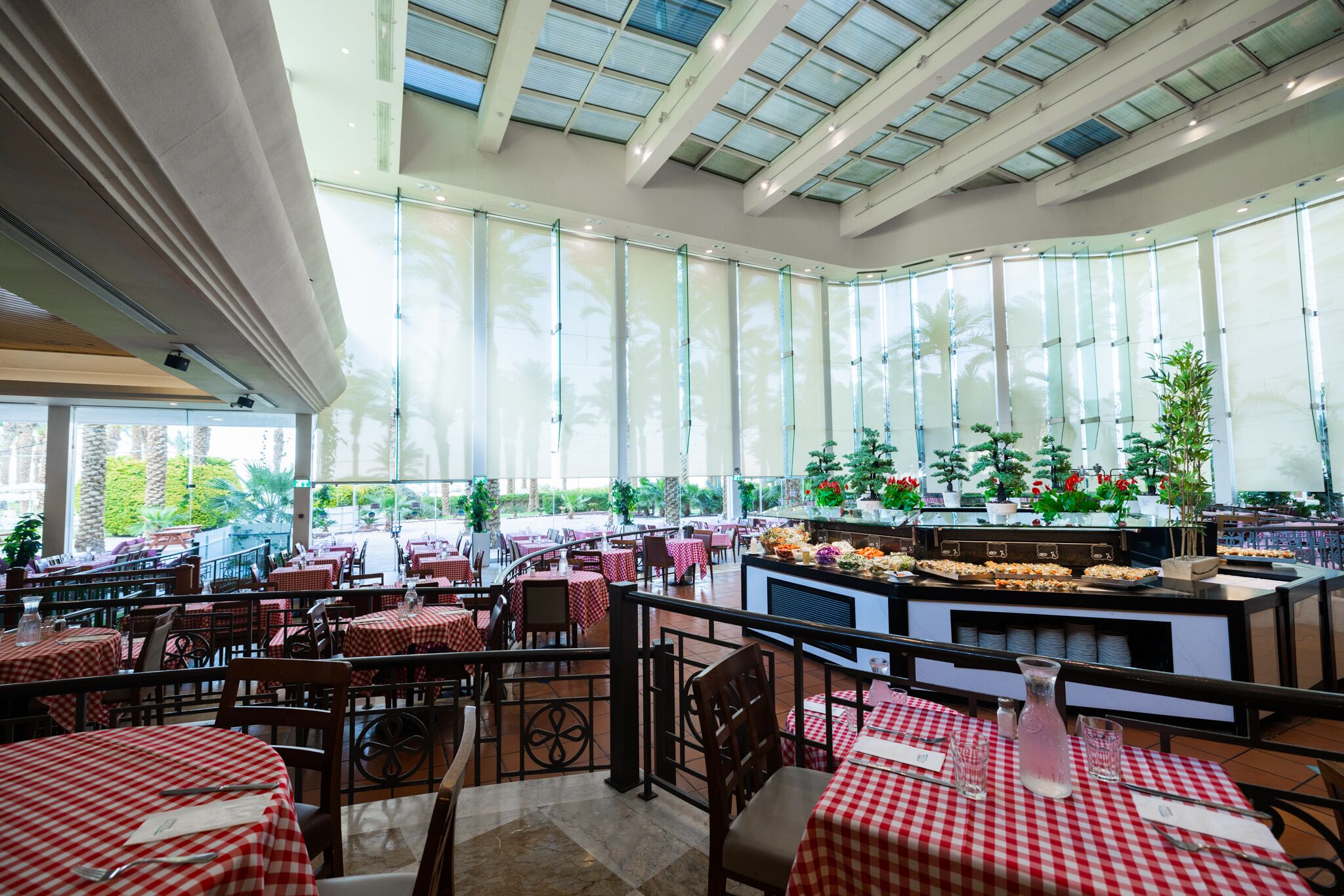 Un restaurante amplio con mesas de cuadros rojos y blancos, una zona de bar central y grandes ventanales con vistas a palmeras y follaje.