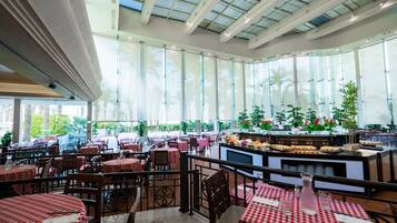 Un restaurante amplio con mesas de cuadros rojos y blancos, una zona de bar central y grandes ventanales con vistas a palmeras y follaje.