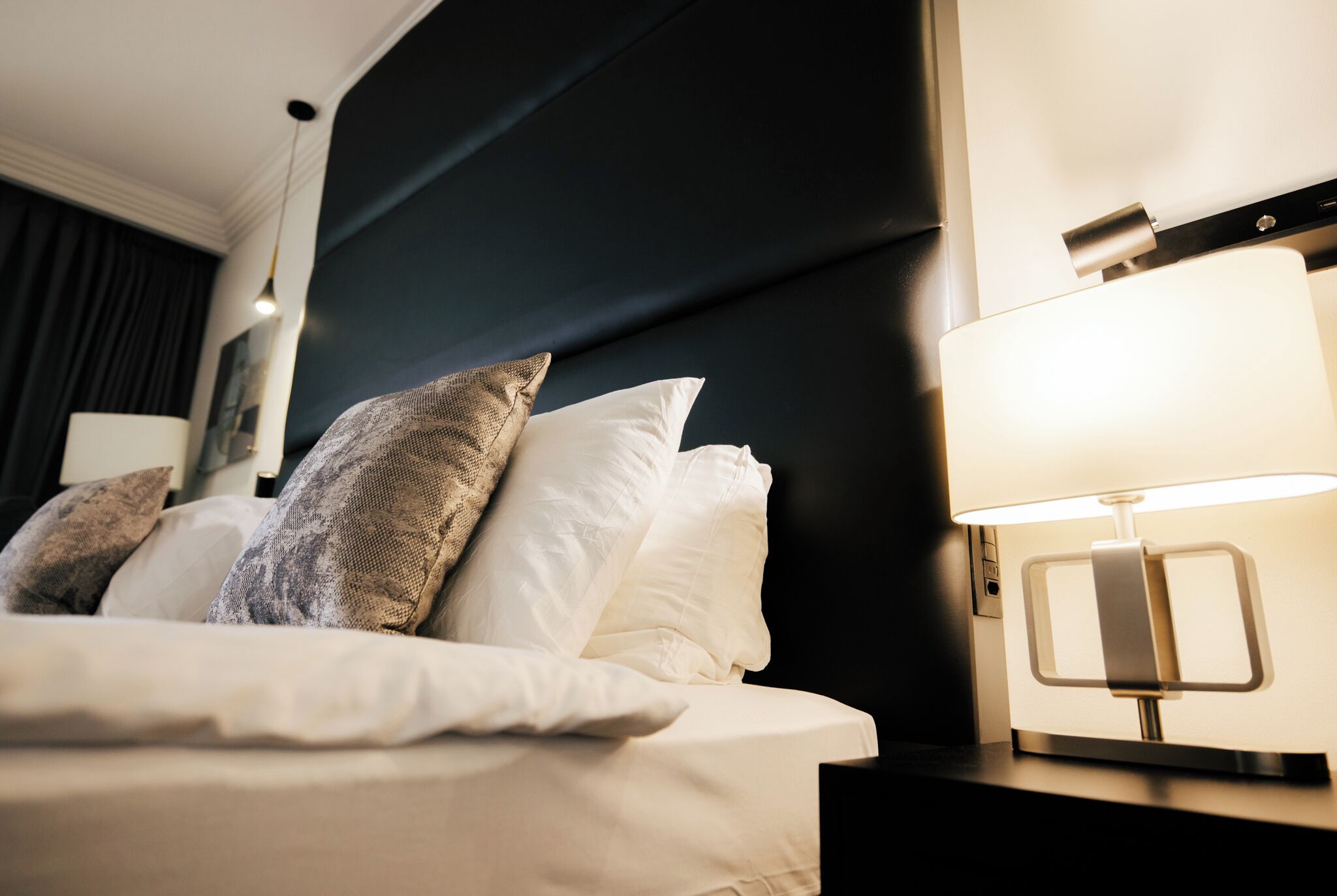 A neatly made bed with white linens and multiple pillows, a modern bedside lamp, and a dark headboard.