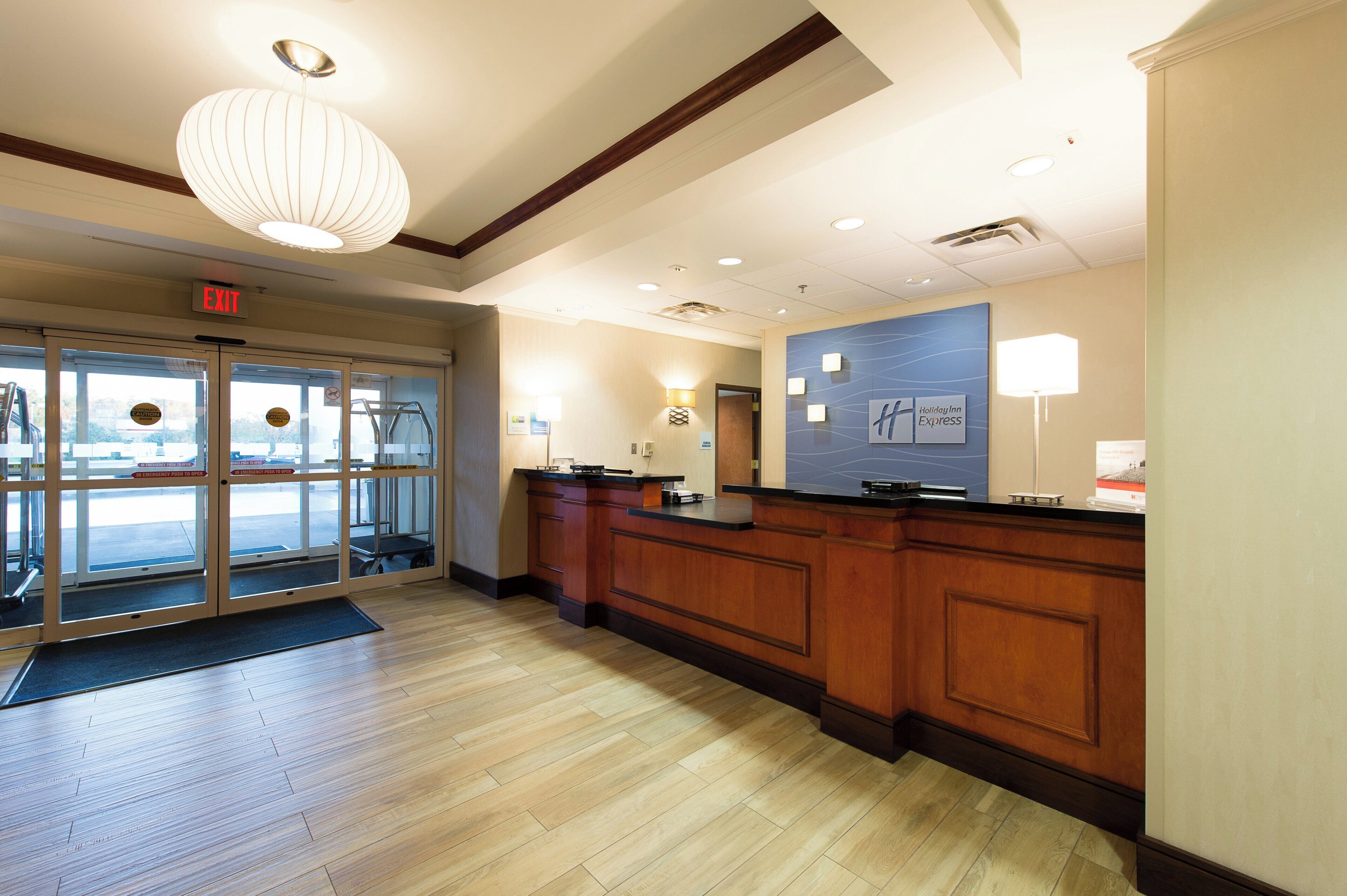 A hotel reception area with a wooden reception desk, a blue wall with the Holiday Inn Express logo, and a glass door entrance.