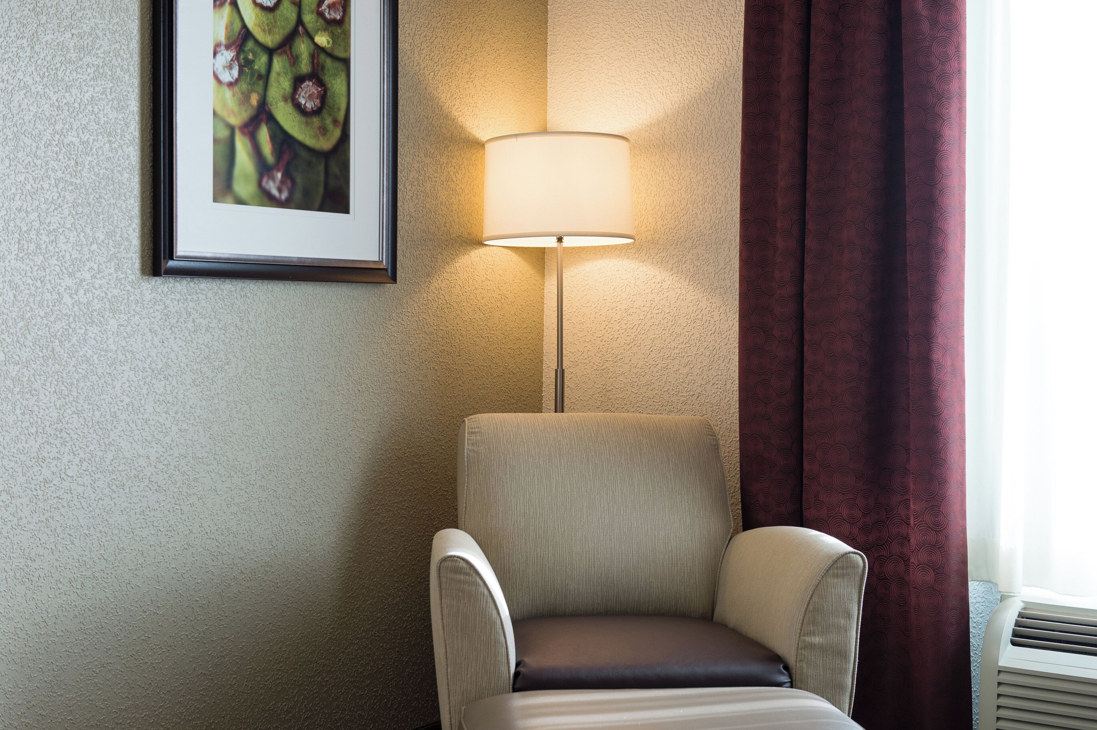 A hotel room with a single armchair, a wall-mounted lamp, and a framed picture.