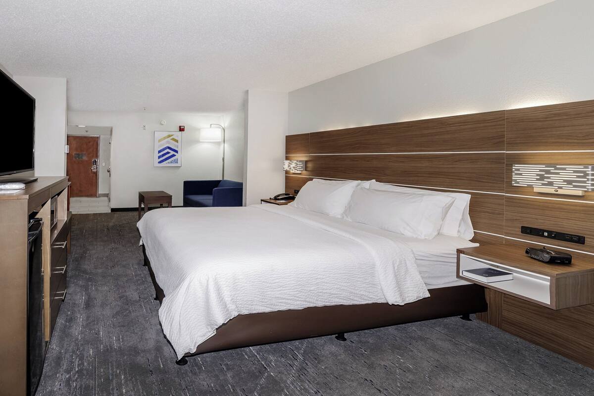 A hotel room with a large bed, a desk with a flat-screen TV, a bedside table, and a wooden headboard.