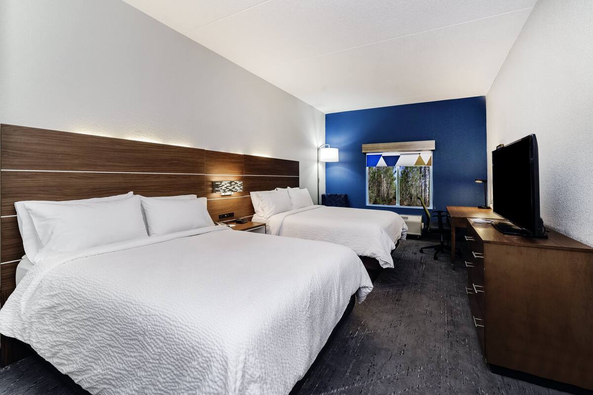 A hotel room with two beds, a desk, a television, and a window with a view of trees.