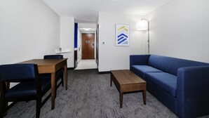 A hotel room with a wooden dining table, blue sofa, and a small wooden coffee table. A hallway leads to the bathroom. - Holiday Inn Exp Walterboro by IHG (Walterboro)