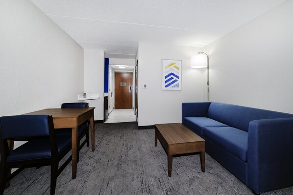 A hotel room with a wooden dining table, blue sofa, and a small wooden coffee table. A hallway leads to the bathroom. - Holiday Inn Exp Walterboro by IHG (Walterboro)