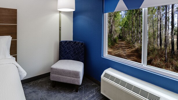 A room with a window view of a forest path, a white air conditioning unit, a grey upholstered bench, and a wall-mounted lamp. - Holiday Inn Exp Walterboro by IHG (Walterboro)