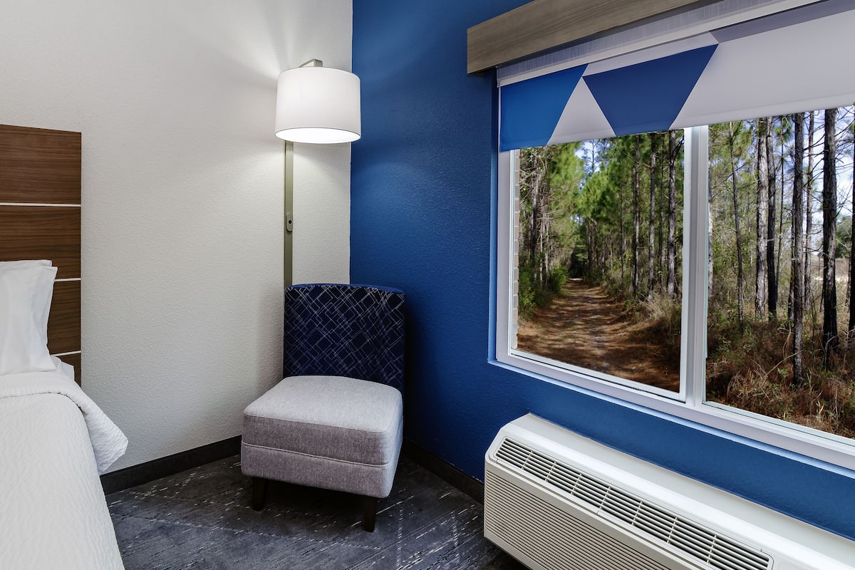 A room with a window view of a forest path, a white air conditioning unit, a grey upholstered bench, and a wall-mounted lamp.