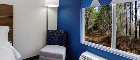 A room with a window view of a forest path, a white air conditioning unit, a grey upholstered bench, and a wall-mounted lamp.