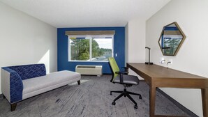 A room with a wooden desk, a green office chair, a grey sofa, and a window with a view of trees and a building. - Holiday Inn Exp Walterboro by IHG (Walterboro)
