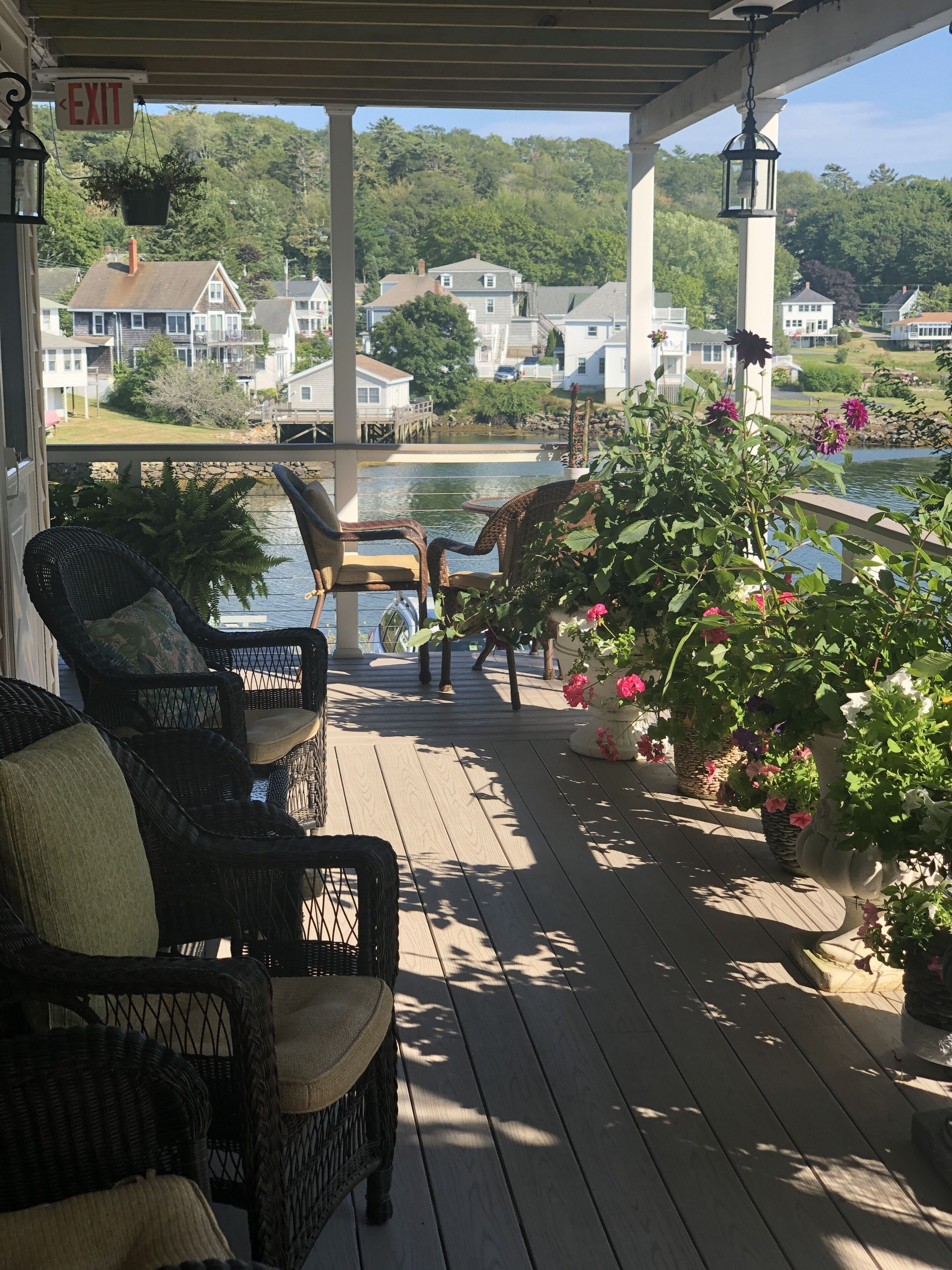 A covered outdoor deck with wicker furniture and potted plants, overlooking a lake with houses in the background.