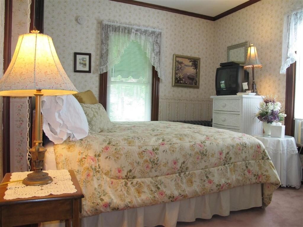 Un dormitorio con cubrecama floral, lámpara de noche, cómoda, televisor y ventana con cortinas de encaje.