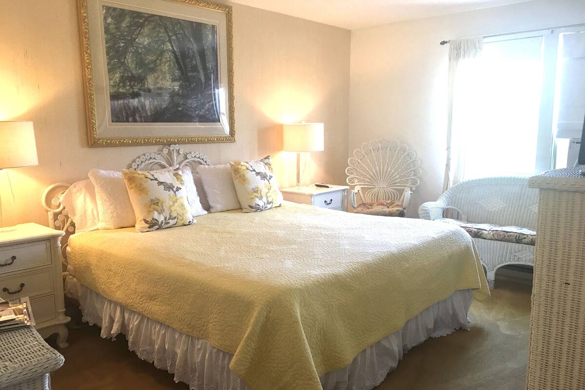 Un dormitorio ordenado con una cama grande, dos mesitas de noche, una silla y un cuadro en la pared.