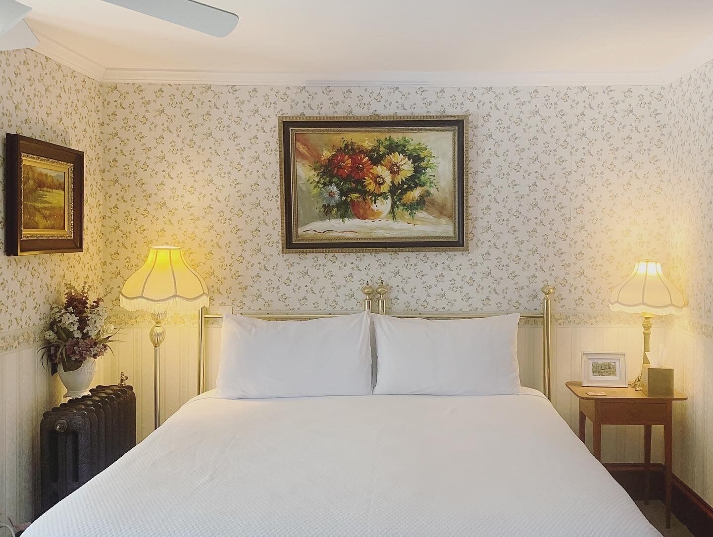 Une chambre d’hôtel avec un grand lit, deux lampes de chevet, un tableau représentant des fleurs au-dessus du lit et un radiateur sur le côté gauche.