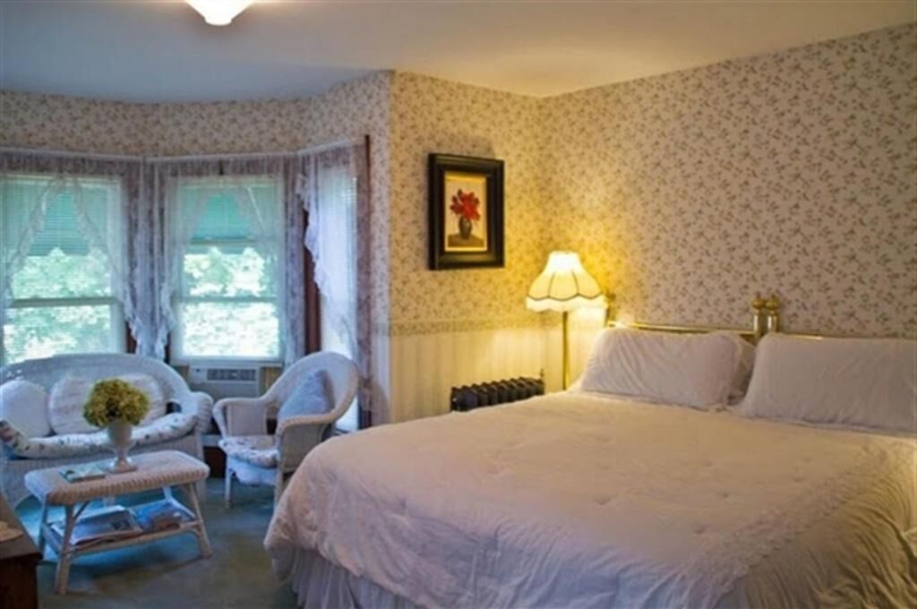 Un dormitorio con una cama grande, dos sillones, una mesa con un jarrón y una lámpara.