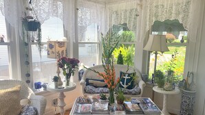 Une pièce chaleureuse, meublée de meubles en osier, avec une table basse sur laquelle sont posés des magazines, et d’où l’on peut admirer le jardin à travers des rideaux en dentelle.