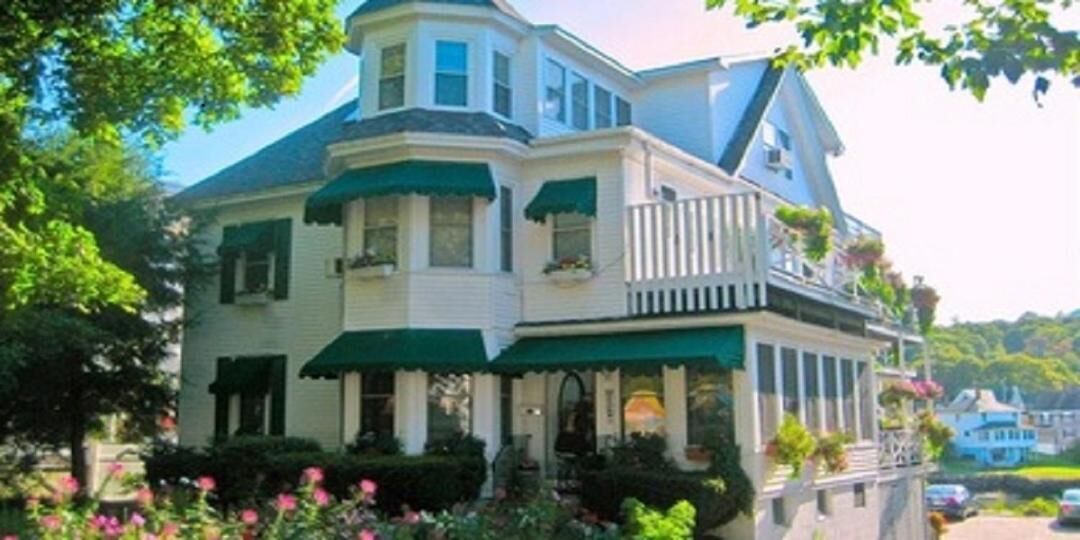 Une grande maison à deux étages, dotée d’une véranda qui fait le tour de la maison, de stores verts et d’un balcon agrémenté de plantes en pot.