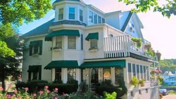 Une grande maison à deux étages, dotée d’une véranda qui fait le tour de la maison, de stores verts et d’un balcon agrémenté de plantes en pot.