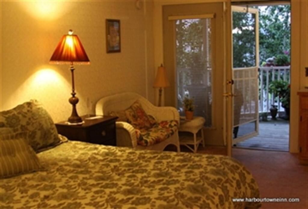 Un dormitorio con cama, mesita de noche, silla, lámpara y puerta corrediza de vidrio que da a un balcón con plantas.