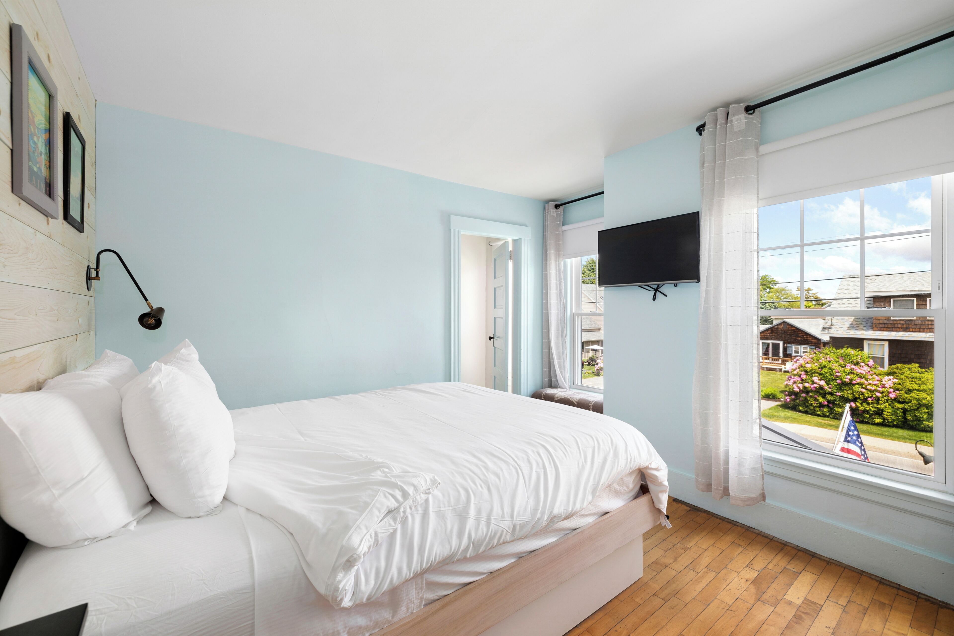 A bedroom with a bed, a mounted TV, a window with curtains, and a view of the outside.