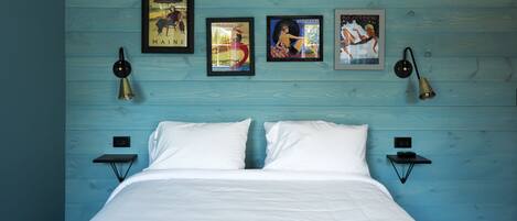 A bed with white linens, two bedside tables, and three framed posters on a blue wooden wall.
