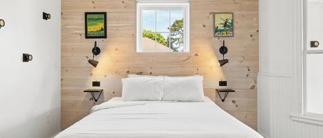 Une chambre à coucher avec une tête de lit en bois, un lit recouvert de linge de lit blanc, deux lampes fixées au mur et deux tableaux encadrés.