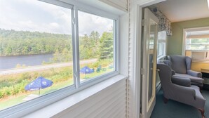 A room with a large window overlooking a lake and forest, a grey armchair, and a small table with a blue umbrella. - Inn By The River (West Forks)